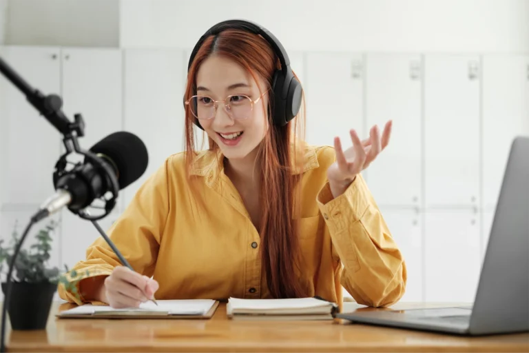 Young woman in yellow shirt with red hair sitting at a desk with a laptop and microphone. She has headphones on and is gesturing as she speaks on GlobalMeet Webcast