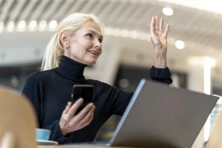An image of a woman sitting at a desk, with a laptop. Her phone is in her hand and she is waving at someone out of frame.